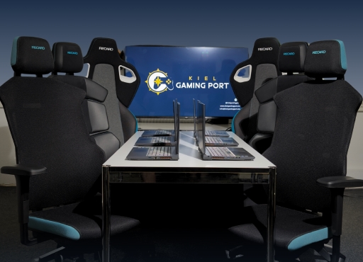 Sechs Recaro Gaming Stühle sind um einen Tisch platziert. Die Sitze sind leicht eingedreht. Auf dem Tisch befinden sich sechs aufgeklappte Laptops.